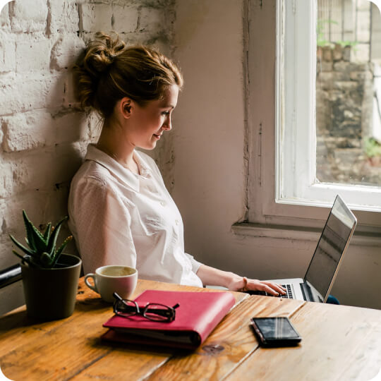 Profile view of a light-skinned person, smiling and using a laptop while seated beside a window