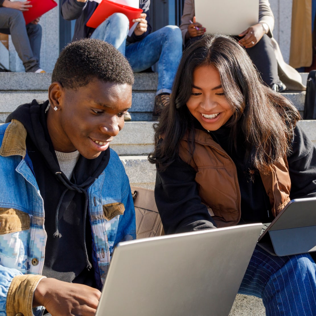 Two students sitting on steps with laptops, studying together outdoors.
