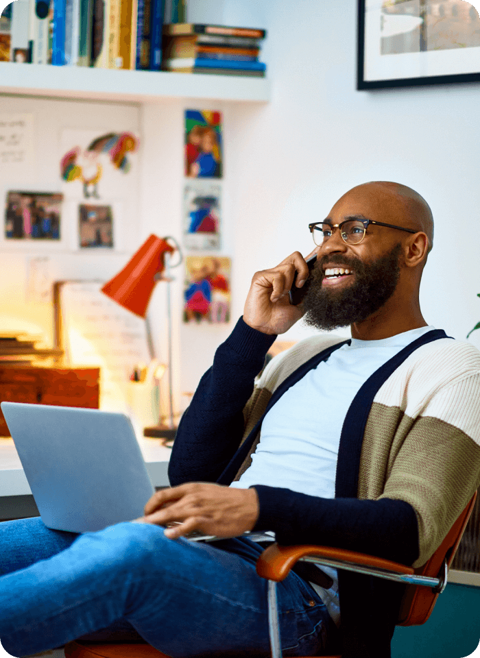 Profile view of a young person with medium dark skin, a beard, and glasses, using a laptop and phone